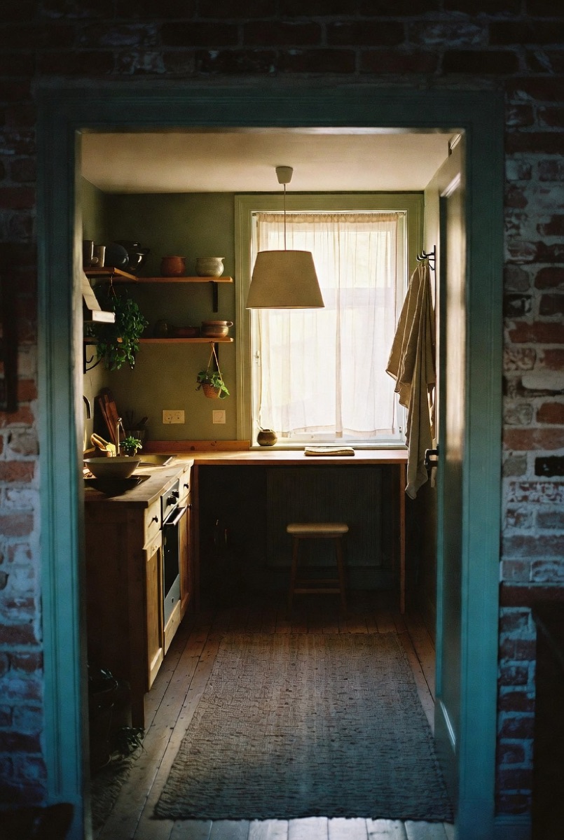 A snug sage-green kitchen with open wood shelves, a pendant lamp and a sheer-curtained window